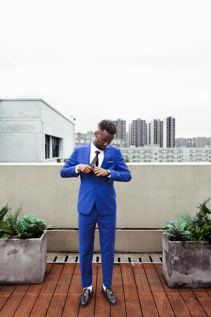 Adult man in blue suit adjusting cuffs on a rooftop terrace with cityscape view.